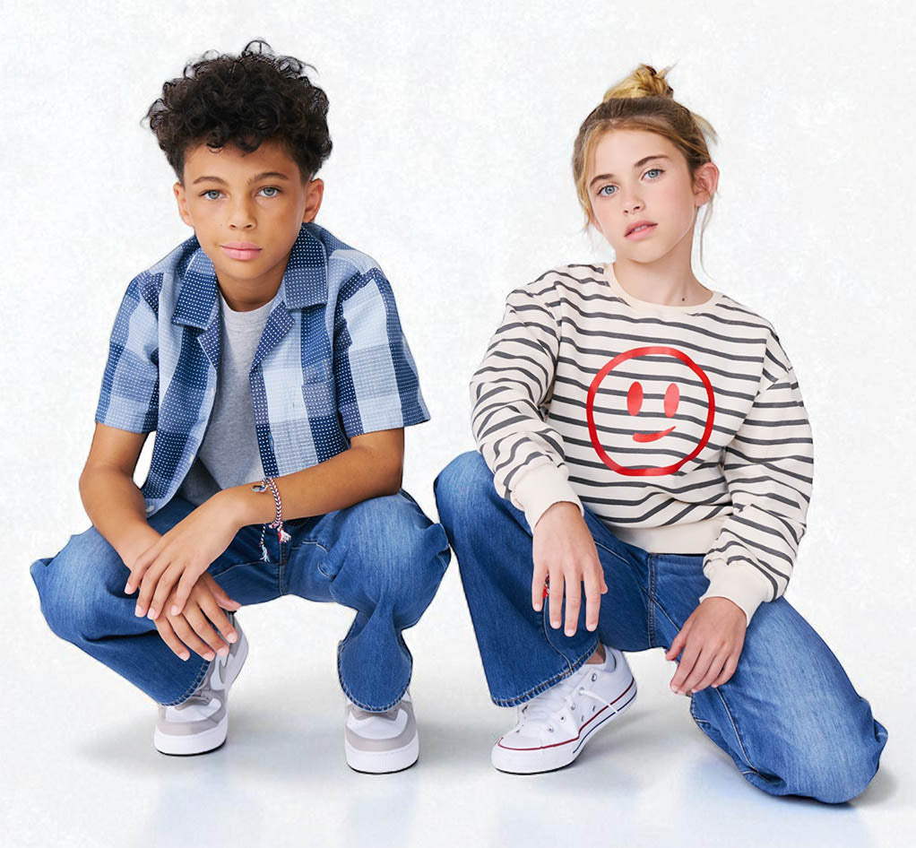 Two children posing against a white background, one wearing a blue plaid shirt and jeans, the other in a striped sweater with a smiley face design.