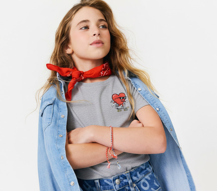 Young girl wearing a red bandana, denim jacket, and gray shirt with a heart on a skateboard  graphic on a white background